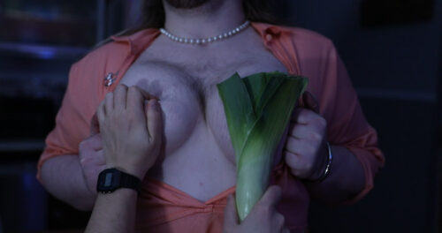 A caucasian person with hairy breasts, with another pair of hands pinching one nipple and holding a fat leek up to the other. This is Aorta Films!