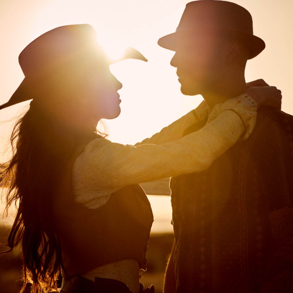 Maya Woulfe and Oliver Davis in the erotic-emotional relationship-driven feature film "California Dreaming" - standing gazing at each other, wearing cowboy and cowgirl wild west gear and cowboy hats, backlit by a setting sun